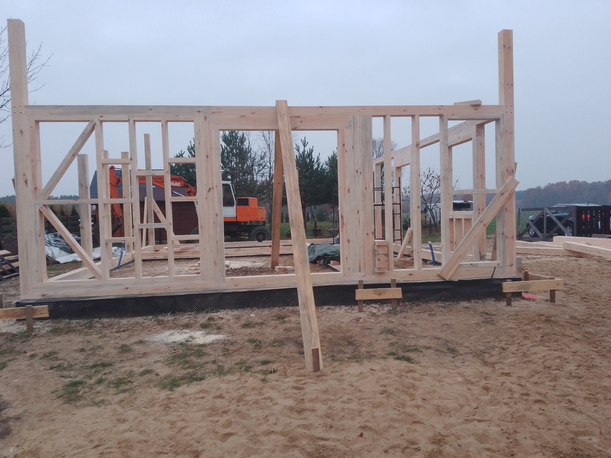 Surowy szkielet domu w trakcie budowy, z widoczną konstrukcją drewnianą i sprzętem budowlanym w tle. Ujęcie z poziomu gruntu, w pochmurny dzień.