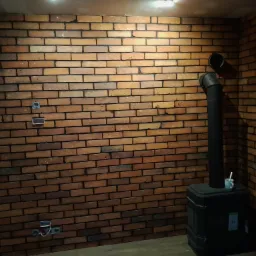 Ściana z cegły klinkierowej wewnątrz pomieszczenia z widocznymi puszkami elektrycznymi i czarnym piecem kominkowym z rurą i kubkiem na górze.