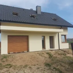 Jednorodzinny dom z beżową elewacją i drewnianą bramą garażową, czarny dach z oknami połaciowymi, widok z zewnątrz na niezagospodarowany teren.