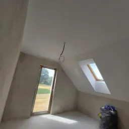 Poddasze w trakcie prac wykończeniowych: ściany pokryte szpachlą, widoczne okno dachowe i okno w ścianie szczytowej, kabel zwisający z sufitu, worek z odpadami budowlanymi w rogu.