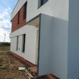 Nowoczesny dom z białą elewacją o fakturze baranka, z elementami wykończenia cegłą klinkierową i grafitowymi detalami wokół okien i wejścia, widoczny fragment przewodu elektrycznego na ziemi.