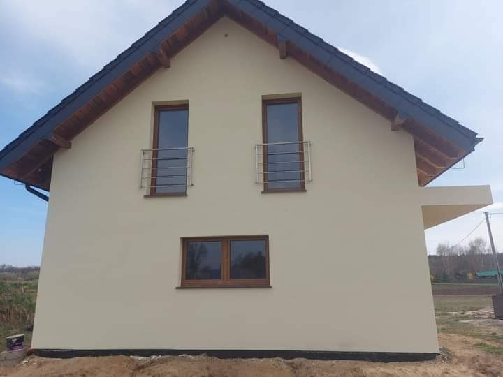 Dwupiętrowy dom jednorodzinny z beżową elewacją, ciemnym dachem i brązowymi oknami z metalowymi balustradami, widoczne zarysowane granice nowej elewacji przy gruncie i puszka farby przy ścianie.