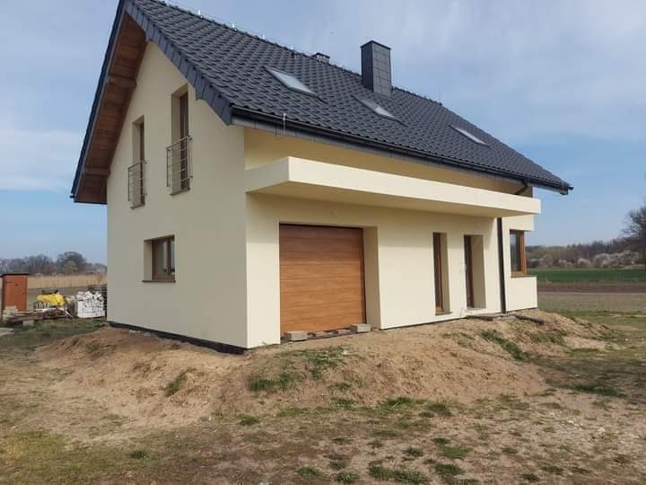 Nowoczesny dom jednorodzinny z jasną elewacją, grafitowym dachem i drewnianą bramą garażową, otoczony nierównym terenem i zielenią w tle.