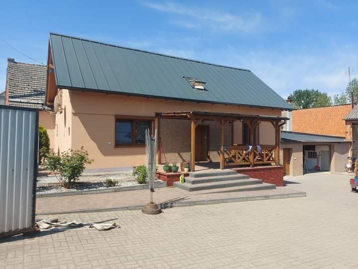 Dom jednorodzinny z beżową elewacją, szarym dachem i drewnianą werandą, widok z brukowanego podjazdu w słoneczny dzień.