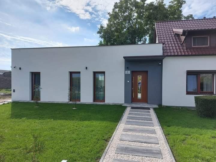 Nowoczesny dom jednorodzinny w Nowym Sielcu z jasną elewacją, brązowymi oknami i drzwiami wejściowymi. Ścieżka z płyt prowadzi do wejścia, otoczona zielonym trawnikiem.