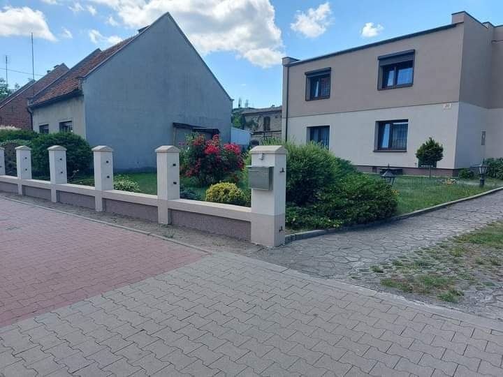 Dwa budynki o różnej architekturze i kolorystyce elewacji, oddzielone niskim, pomalowanym płotem z betonowymi słupkami, widoczne z poziomu chodnika z kostki brukowej.