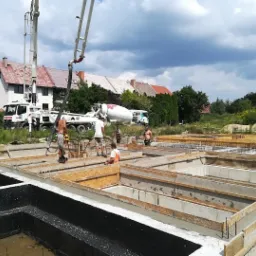 Wylewanie betonu do szalunków fundamentowych pod budowę domu jednorodzinnego; widoczne pompy do betonu, betoniarka i pracownicy w trakcie pracy.