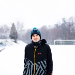 Uśmiechnięty mężczyzna w sportowej kurtce i czapce na tle zasypanego śniegiem boiska.