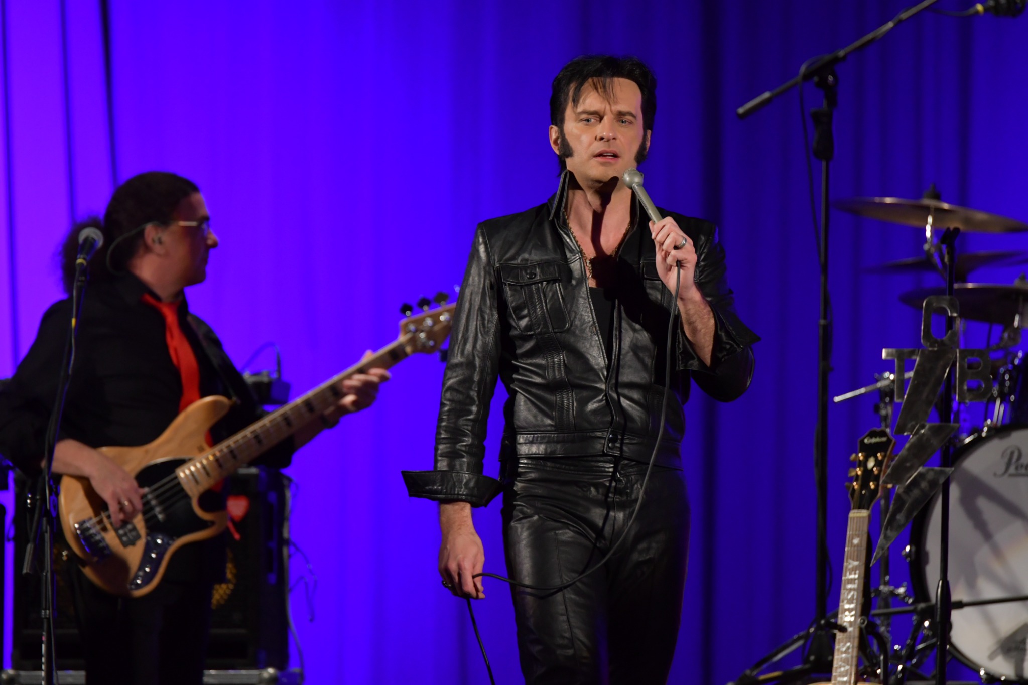 Wokalista w czarnym skórzanym kombinezonie śpiewa do mikrofonu na scenie z basistą w tle i perkusją po prawej stronie, gitara z napisem 'Elvis Presley' oparta o statyw.