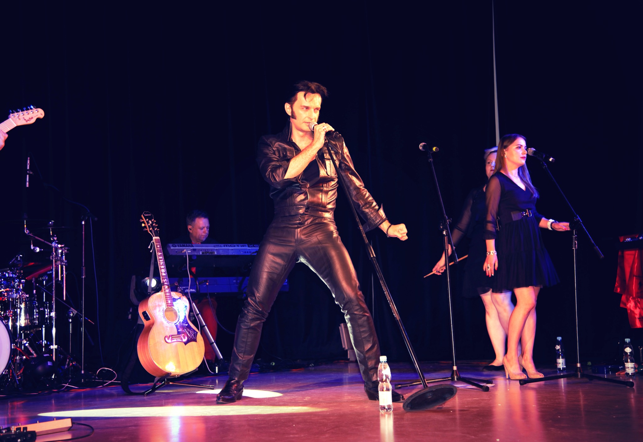 Wokalista w czarnym, skórzanym stroju na scenie, w tle zespół i instrumenty, butelka wody na podłodze.