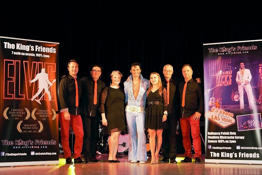 Zespół muzyczny The King's Friends na scenie, z wokalistą przebranym za Elvisa Presleya, otoczony przez pozostałych członków zespołu w czarnych koszulach i czerwonych krawatach i spodniach, obok...