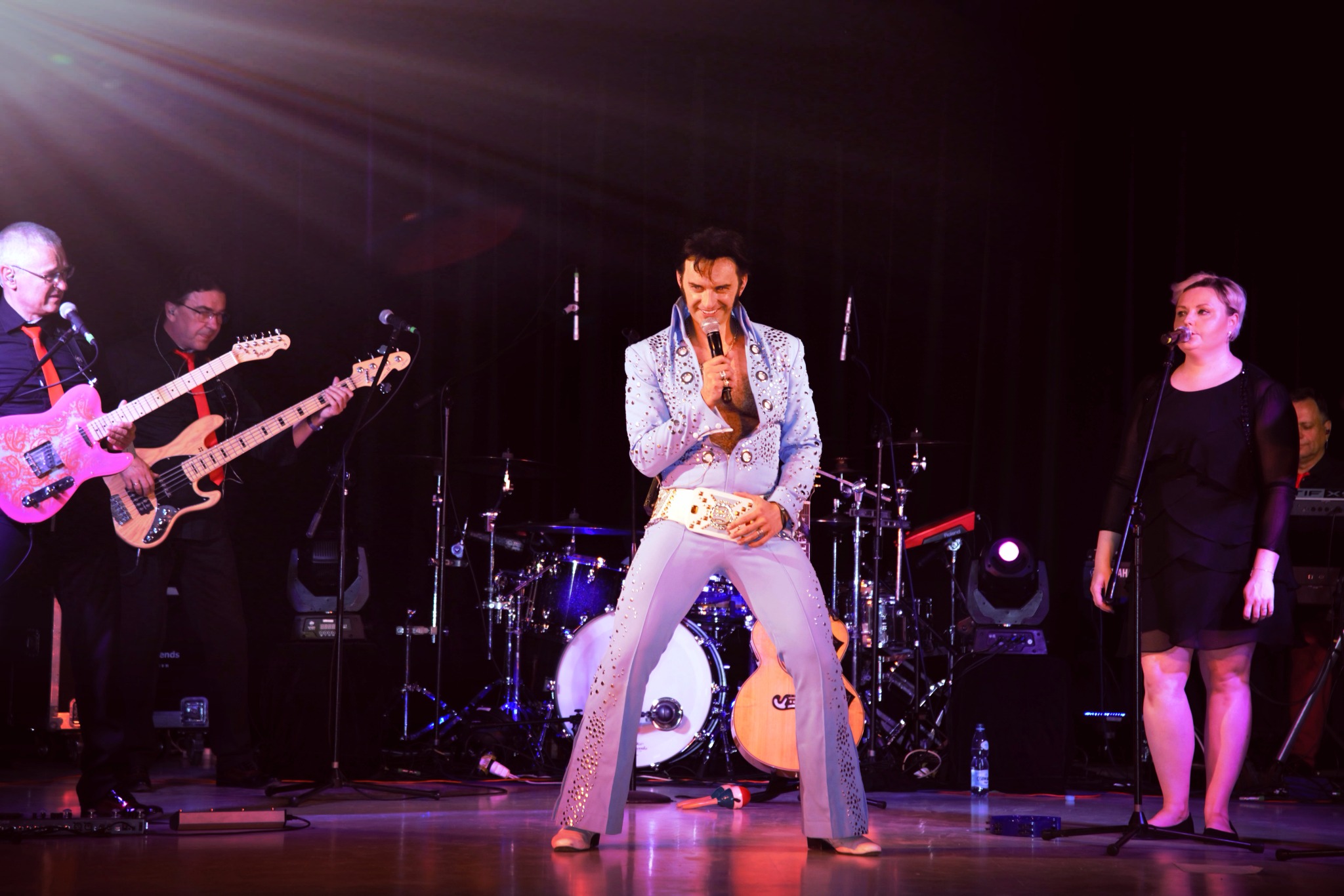 Wokalista w błękitnym kostiumie w stylu Elvisa Presleya z mikrofonem w dłoni na scenie z zespołem muzycznym w tle, oświetlonym reflektorami.