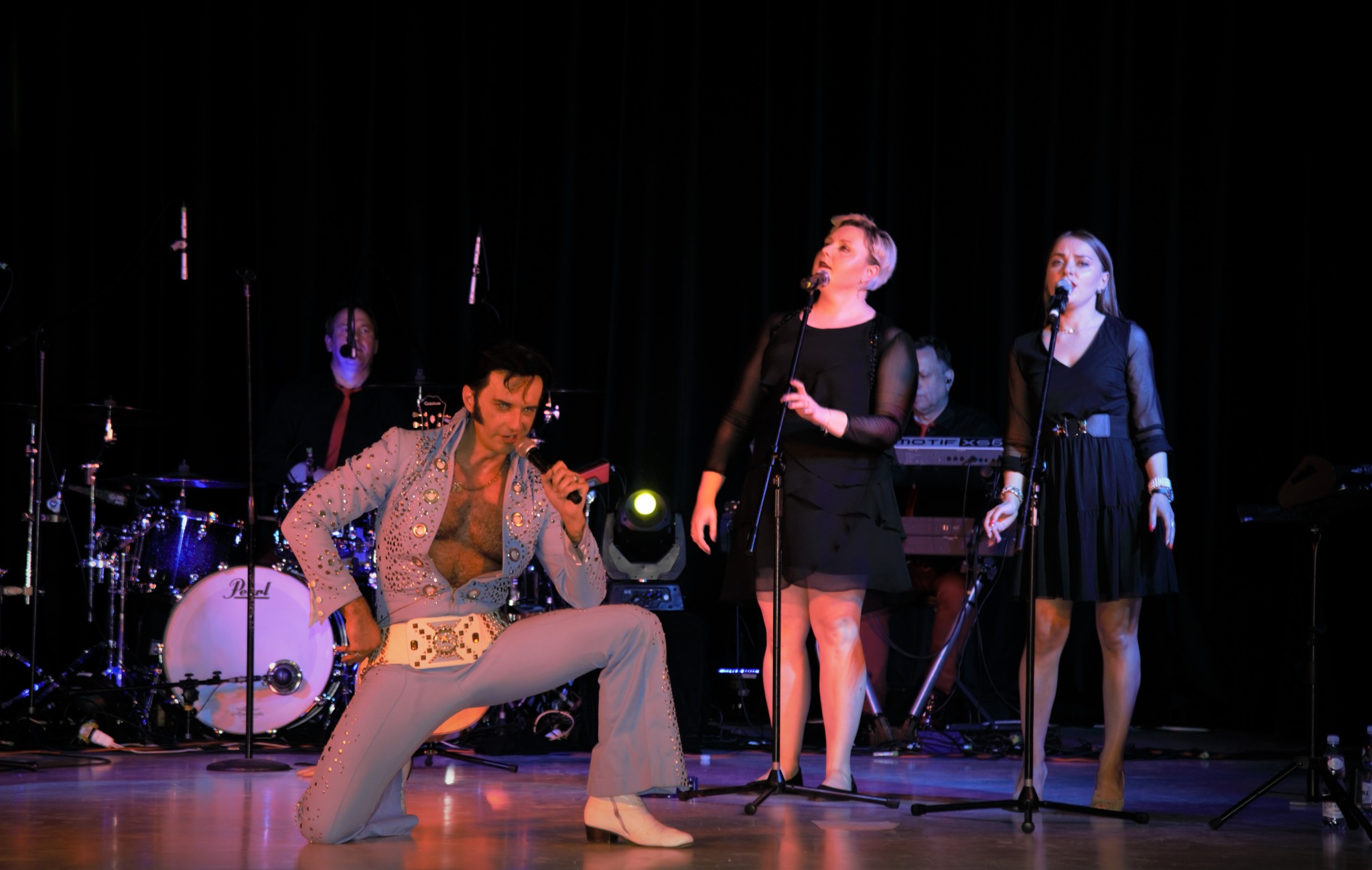 Występ zespołu muzycznego w Warszawie: wokalista w kostiumie Elvisa Presleya klęczy na scenie z mikrofonem, obok dwie wokalistki w czarnych sukienkach, w tle perkusja i klawiszowiec.