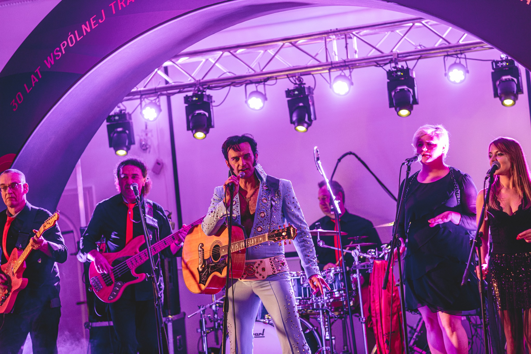 Zespół muzyczny w trakcie występu na scenie, wokalista w kostiumie Elvisa Presleya gra na gitarze, w tle perkusja i wokalistki, oświetlenie sceniczne w odcieniach fioletu.