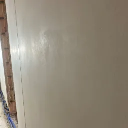 Świeżo nałożony tynk na ścianie wewnętrznej, widoczne ślady po narzędziach, z boku fragment muru z cegieł i niebieskie rurki.