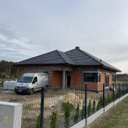 Surowy stan domu jednorodzinnego z czerwoną cegłą, czarną dachówką i częściowo ocieplonymi ścianami, zaparkowany srebrny van z reklamą firmy, ogrodzenie panelowe z podmurówką betonową i młodymi...