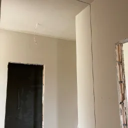 Wnętrze w trakcie prac wykończeniowych: ściany po nałożeniu tynku, widoczne przewody elektryczne, surowy otwór drzwiowy.