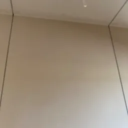 Świeżo pomalowany narożnik pokoju w beżowym odcieniu, widoczne delikatne linie łączenia ścian i sufitu, pojedynczy ślad po taśmie malarskiej na suficie.