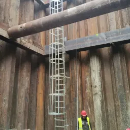 Głęboki wykop zabezpieczony stalowymi ściankami Larsena, z metalową klatką schodową i pracownikami w kaskach i kamizelkach odblaskowych na dnie, obok sterty prętów zbrojeniowych.