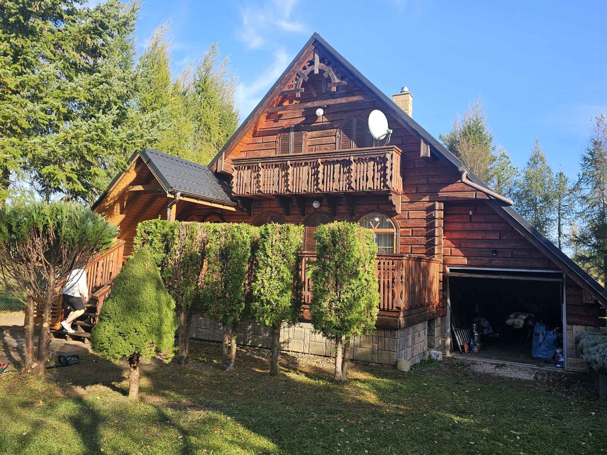 Drewniany dom z balkonem i garażem, otoczony zielenią. Osoba wchodzi po schodach. W tle błękitne niebo. Widoczne drzewa i krzewy.