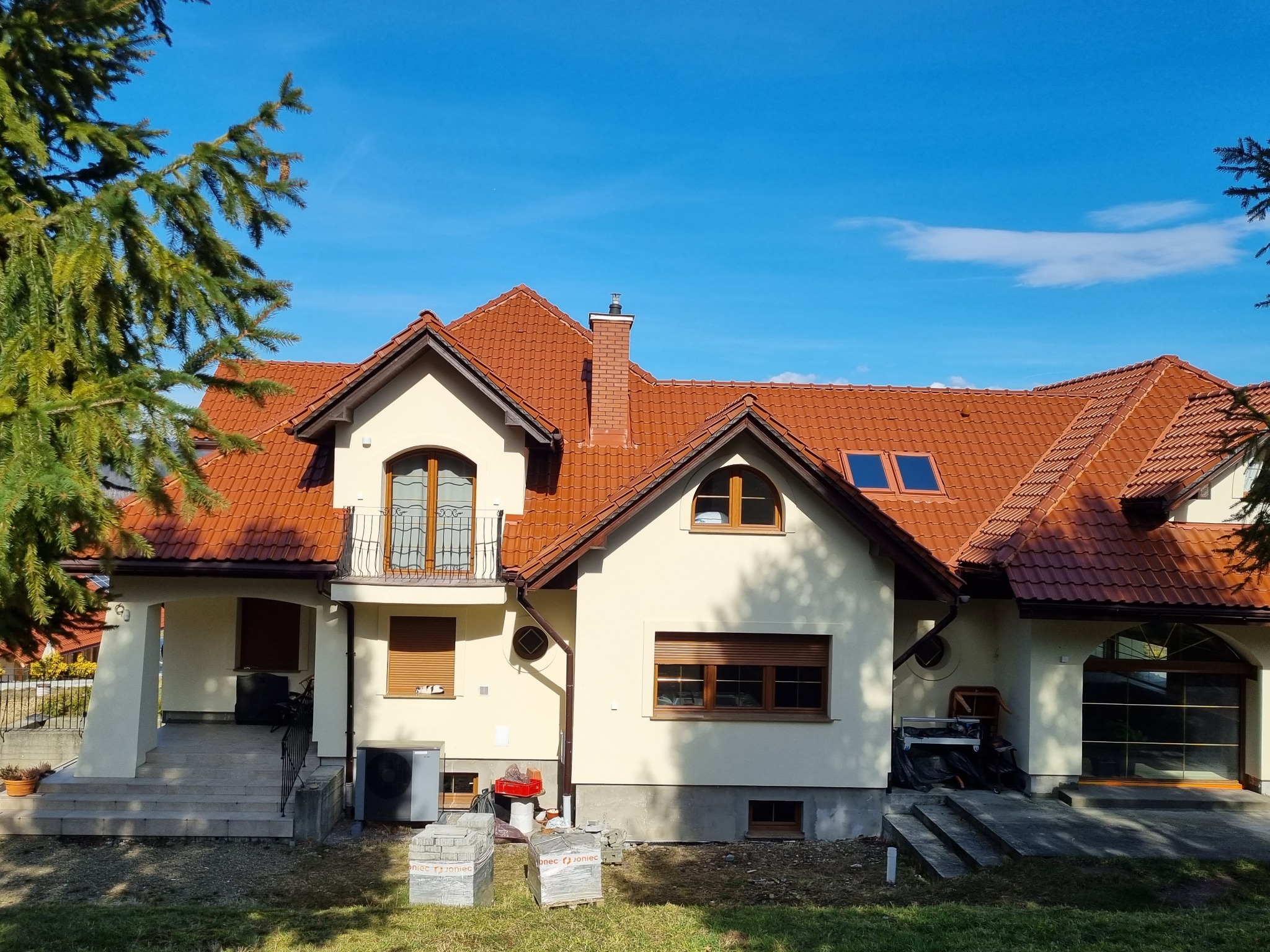 Jasna elewacja domu z czerwoną dachówką w słoneczny dzień. Na pierwszym planie kostka brukowa, sugerująca prace wykończeniowe.