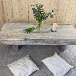 Niski, prostokątny stolik kawowy z surowego, bielonego drewna, ustawiony na szarym dywanie z dwoma białymi poduszkami pod spodem, udekorowany bukietem w szklanym wazonie, świecą i kilkoma tulipanami.