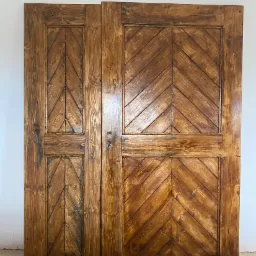 Dwa skrzydła brązowych, drewnianych drzwi z wzorem jodełki, o wyraźnym usłojeniu i widocznych śladach ręcznej obróbki, opartych o białą ścianę i fragment listwy przypodłogowej.