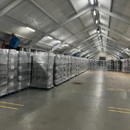 Rząd palet z zapakowanymi towarami w folii stretch w hali magazynowej z szarym betonowym podłożem i żółtymi liniami wyznaczającymi strefy.