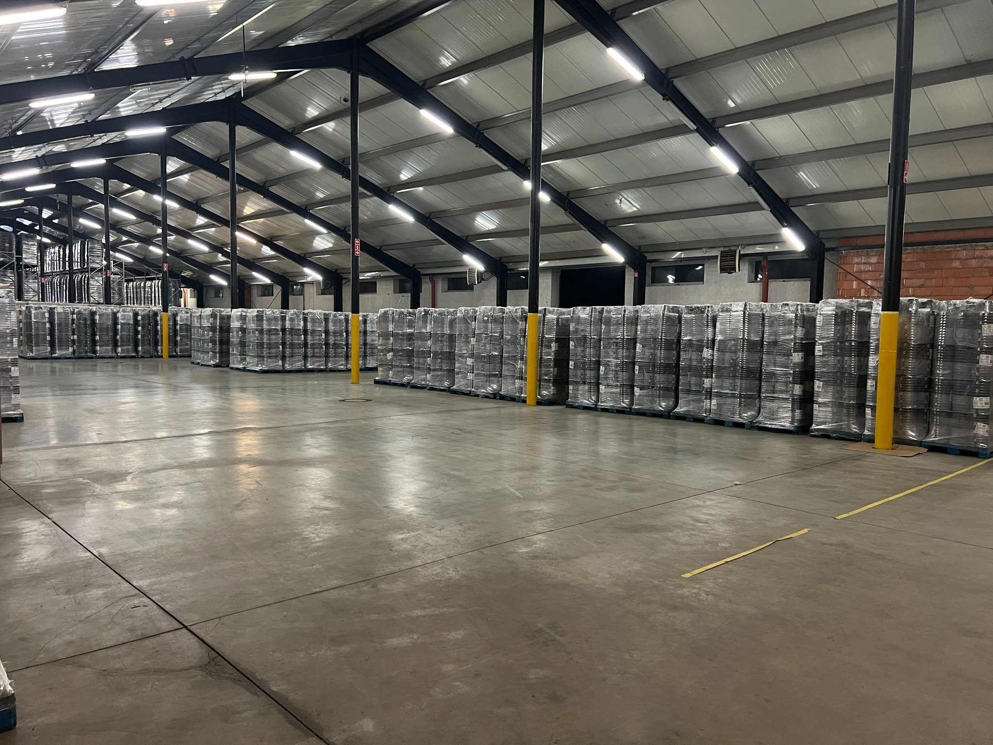 Pełny magazyn z paletami owiniętymi folią stretch, oświetlony szeregiem lamp LED pod metalową konstrukcją dachu.