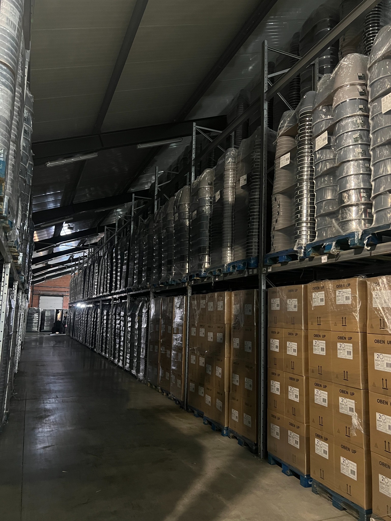 Wnętrze magazynu z regałami wypełnionymi paletami z tekturowymi pudłami oznaczonymi 'OBEN UP' oraz ułożonymi w stosy metalowymi elementami zapakowanymi w folię, widok w głąb magazynu.