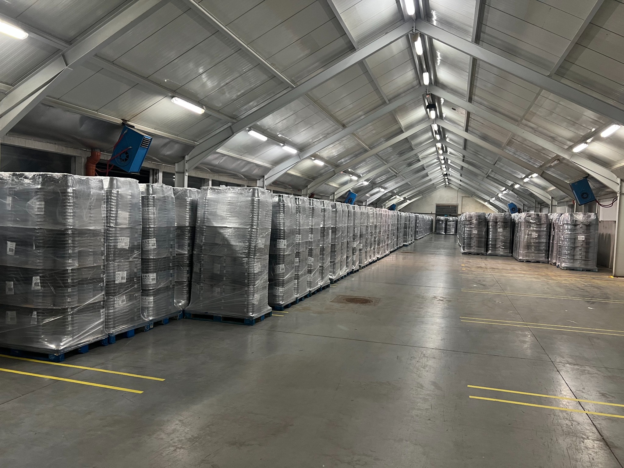 Rząd palet z zapakowanymi towarami w folii stretch w hali magazynowej z szarym betonowym podłożem i żółtymi liniami wyznaczającymi strefy.