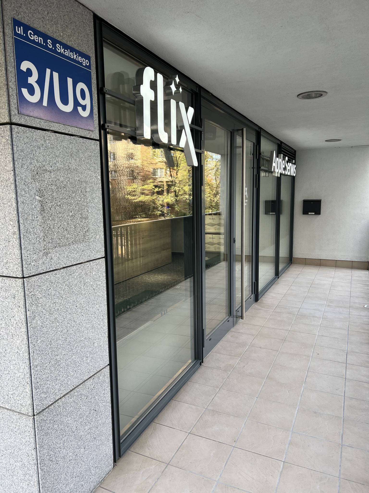 Wejście do serwisu Flix Apple Serwis z widocznym adresem ul. Gen. S. Skalskiego 3/U9, przeszklone drzwi odbijają drzewa.