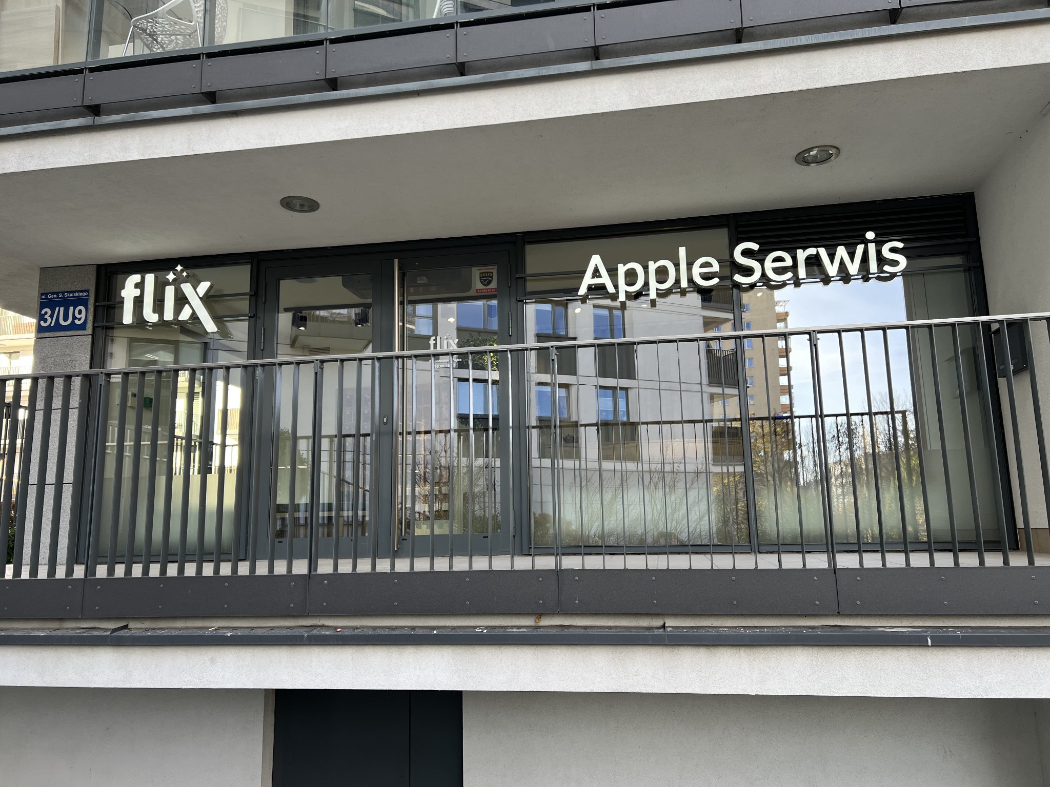 Zewnętrzny widok balkonu lokalu usługowego z napisem 'flix' oraz 'Apple Serwis' na szybach, widoczny numer lokalu 3/U9
