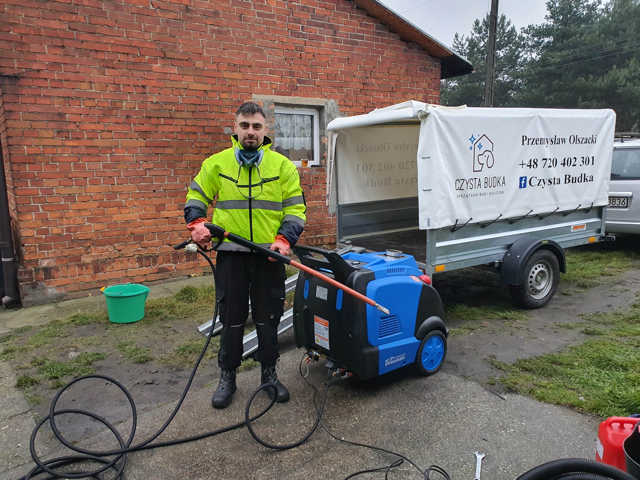 Mężczyzna w odblaskowej kurtce prezentuje niebieski parowy czyszczak ciśnieniowy na przyczepie z reklamą firmy sprzątającej budy i kojce, stojąc na tle ceglanego budynku.
