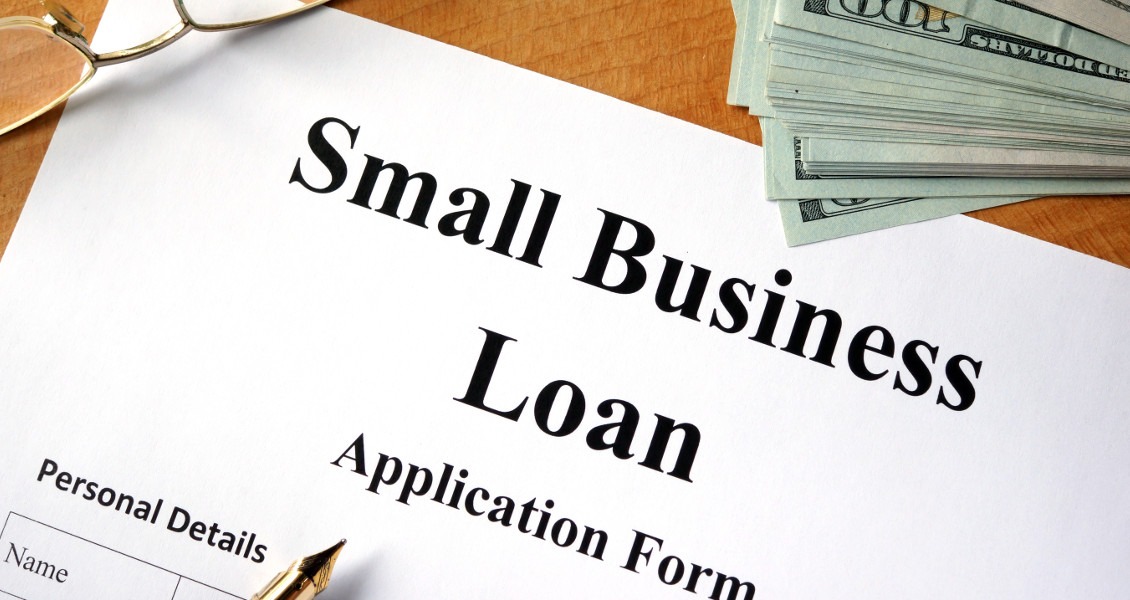 Formularz wniosku o kredyt dla małych firm z piórem wiecznym i stosem studolarowych banknotów na drewnianym biurku, okulary w rogu.