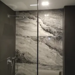 Szklana kabina prysznicowa z deszczownicą, ściany wyłożone wielkoformatowymi płytami imitującymi marmur, widoczne niedokończone prace wykończeniowe.