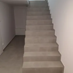 Wykończone schody wewnętrzne z szarych płytek, prowadzące do góry, widoczne płytki podłogowe w tym samym kolorze, białe ściany i drzwi.