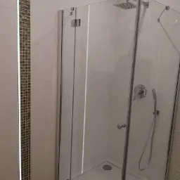 Nowoczesna kabina prysznicowa z chromowanymi okuciami i deszczownicą, obok pionowy pas mozaiki z podświetleniem LED na jasnych płytkach.