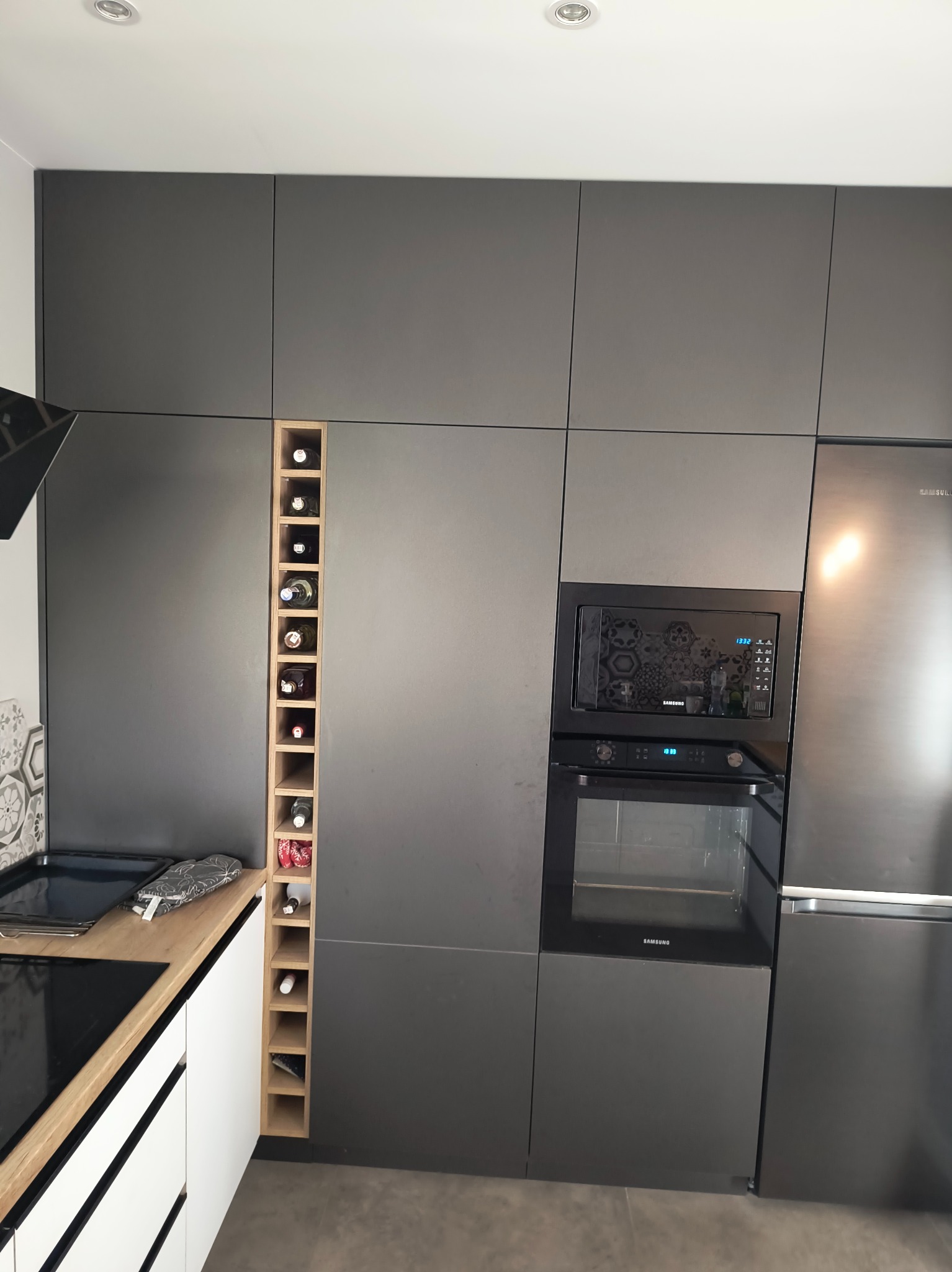 Szara zabudowa kuchenna z drewnianym regałem na wino, wnęką z piekarnikiem i mikrofalą oraz lodówką. Minimalistyczny design, nowoczesne wykończenie.