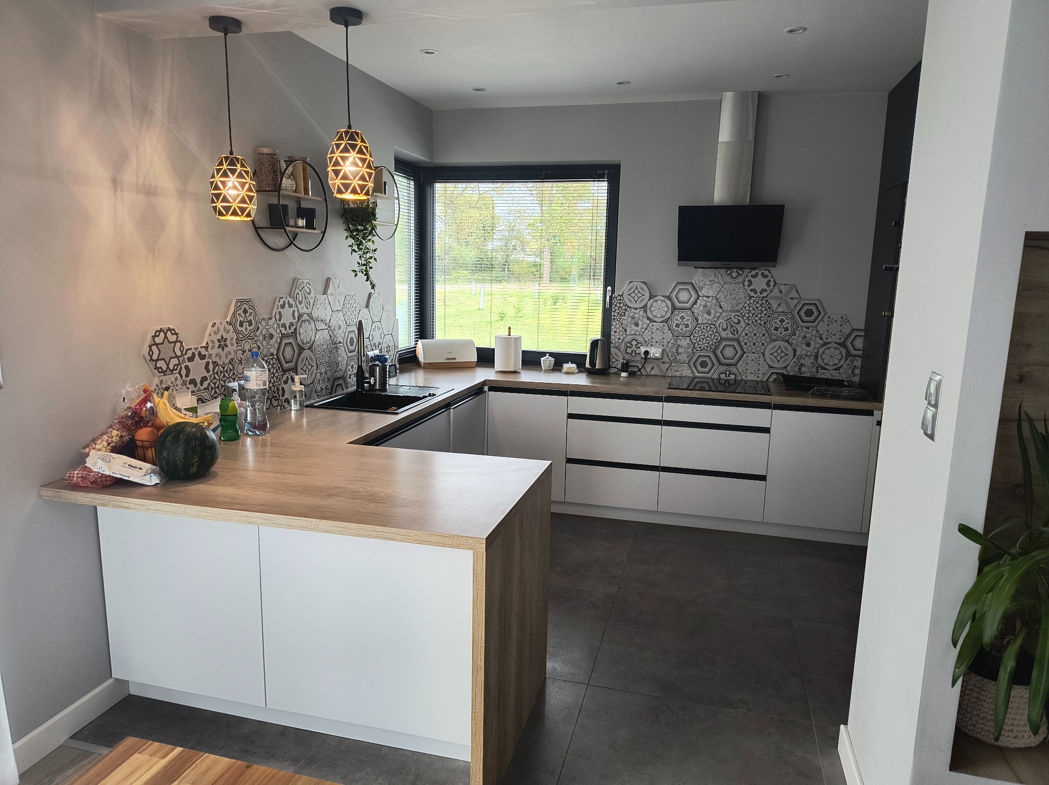 Nowoczesna, jasna kuchnia z drewnianym blatem, heksagonalnymi płytkami nad blatem i lampami wiszącymi. Widok na okno i ogród.