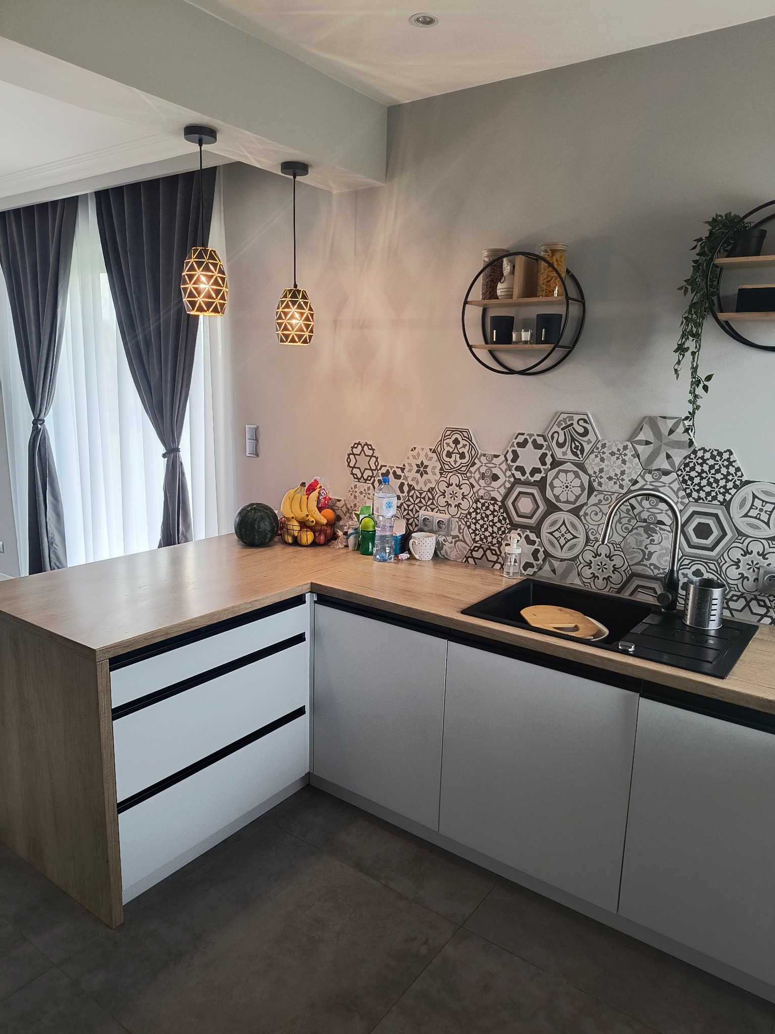 Nowoczesna kuchnia z drewnianym blatem, białymi frontami szafek i heksagonalnymi płytkami nad zlewem. Widoczne oświetlenie i dekoracje ścienne.