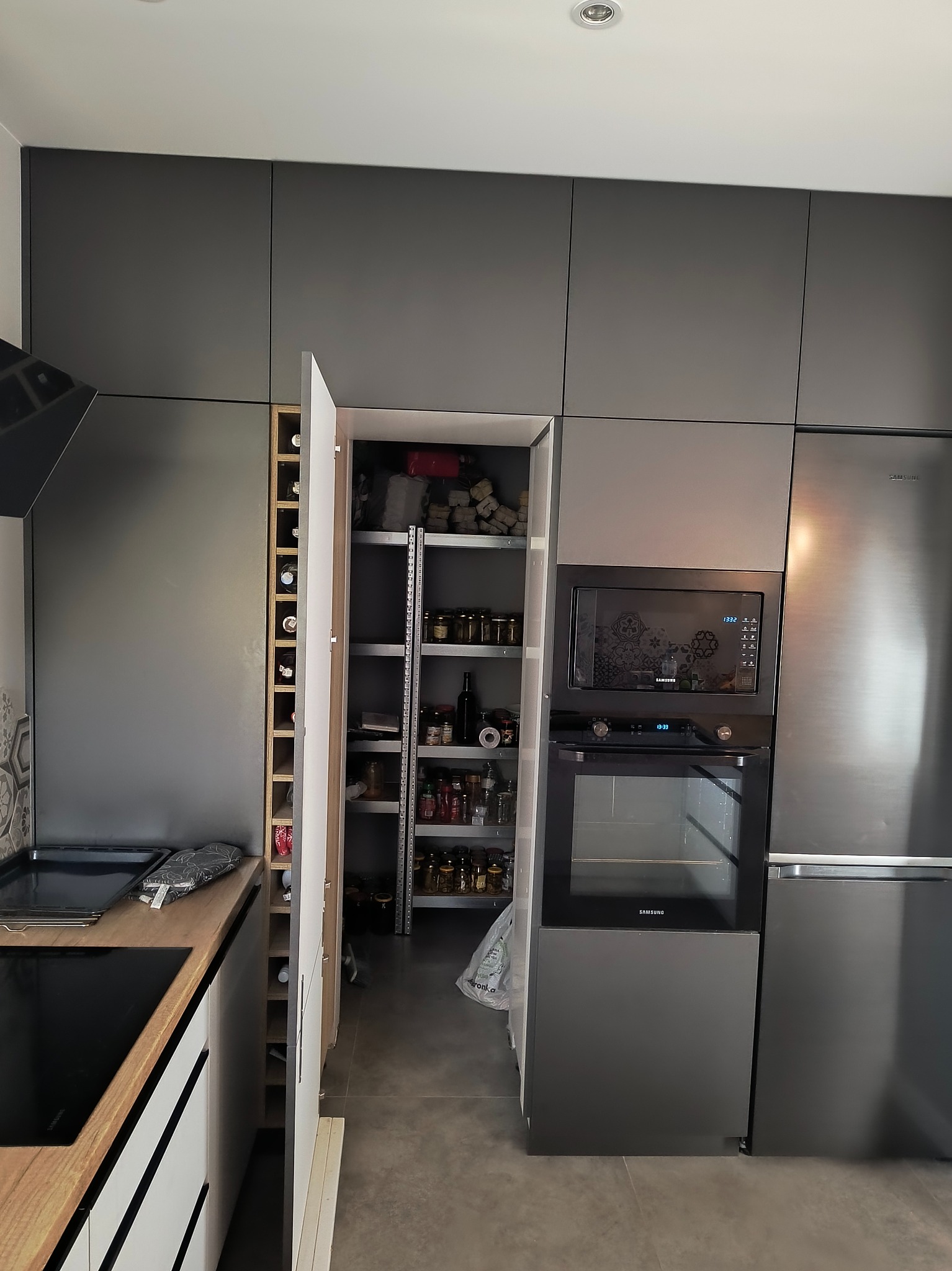 Szara kuchnia z szafkami na wymiar, otwartą spiżarnią i wbudowanym sprzętem AGD. Widoczny regał na wino. Funkcjonalne rozwiązanie do przechowywania w kuchni.