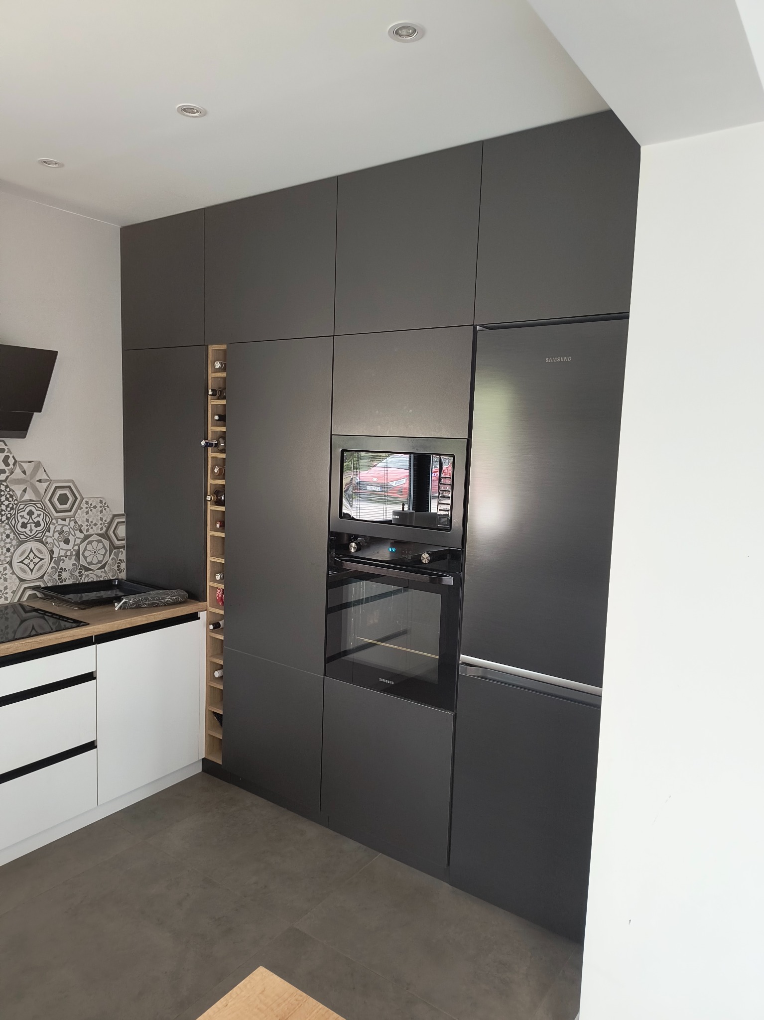 Nowoczesna, grafitowa kuchnia na wymiar z wbudowanym sprzętem AGD Samsung i regałem na wino, uzupełniona białymi szafkami i płytkami w stylu retro.