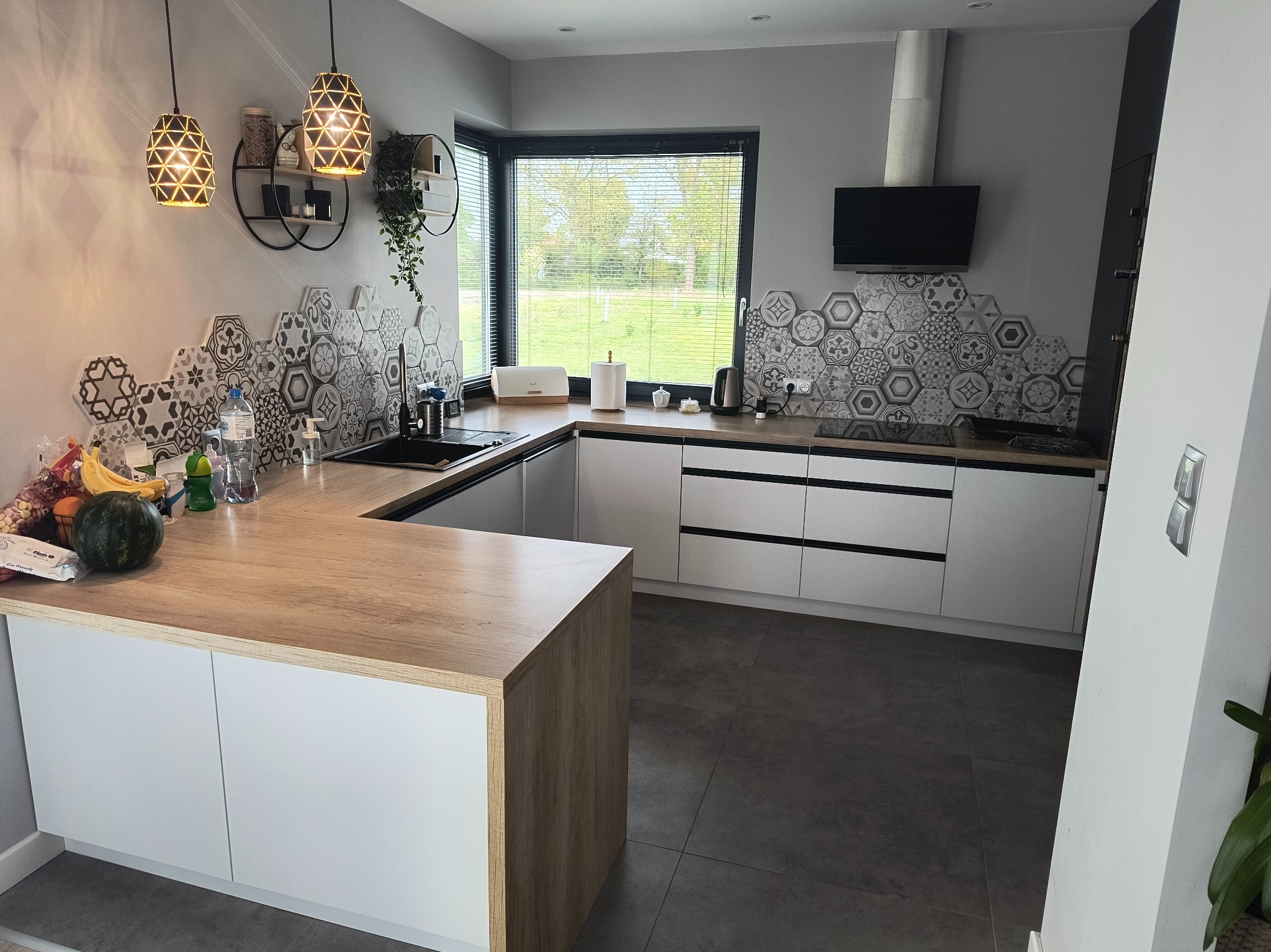 Jasna kuchnia w stylu nowoczesnym z drewnianym blatem i heksagonalnymi płytkami nad blatem. Widoczne oświetlenie i okno z widokiem na zieleń.