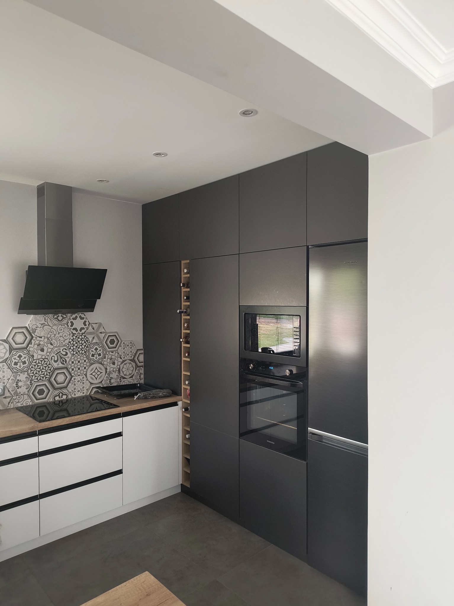 Nowoczesna szara kuchnia z białymi dolnymi szafkami i drewnianym blatem. Uwagę przykuwają heksagonalne płytki nad indukcją i wnęka na wino w wysokiej zabudowie.