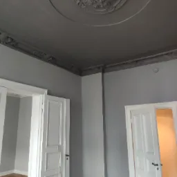 Pokój po remoncie z szarymi ścianami, białymi drzwiami i ciemnoszarym, ozdobnym sufitem z rozetą.