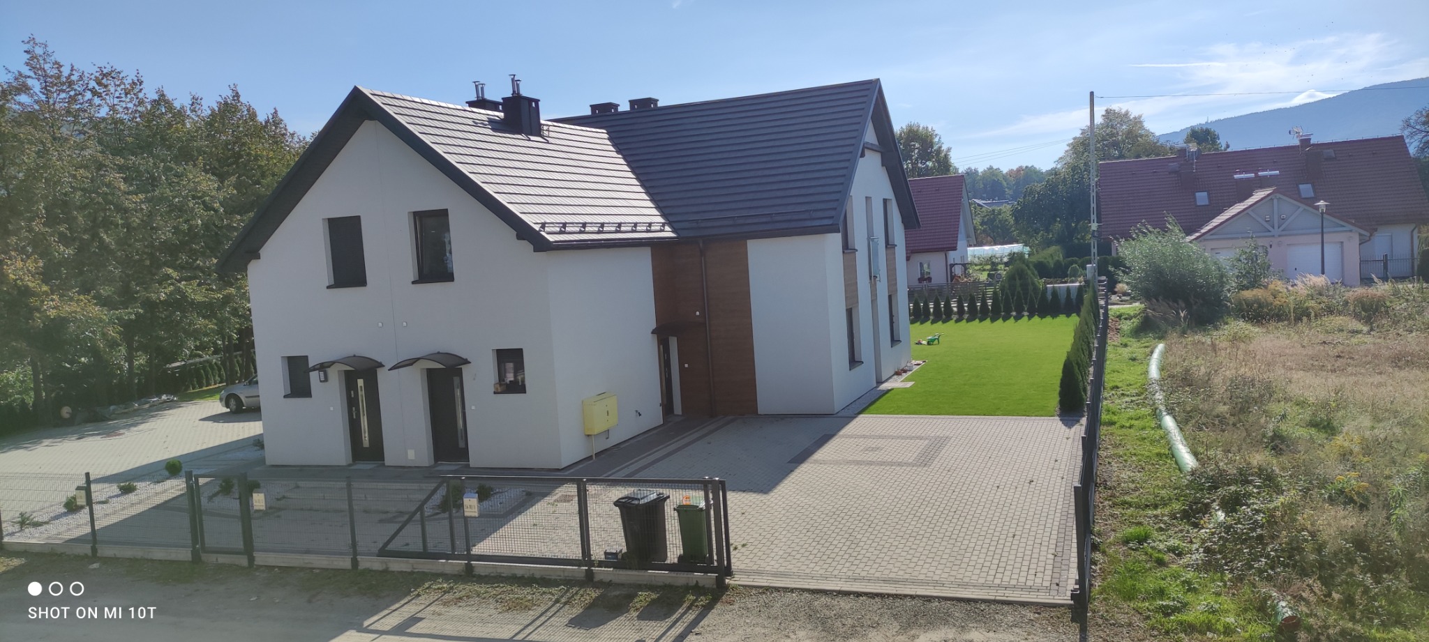 Nowoczesny dom jednorodzinny z jasną elewacją, ciemną dachówką i drewnianymi akcentami, widoczny z zewnątrz, z zadbanym trawnikiem i kostką brukową na podjeździe.