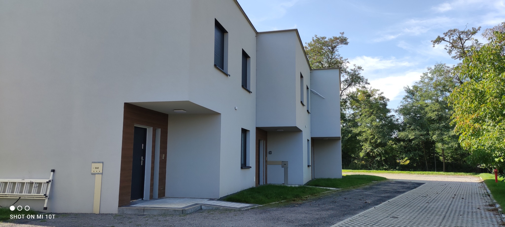 Nowoczesne segmenty mieszkalne z białymi elewacjami i drewnianymi akcentami przy wejściach, widoczne z zewnątrz, z aluminiową drabiną opartą o ścianę i skrzynką gazową