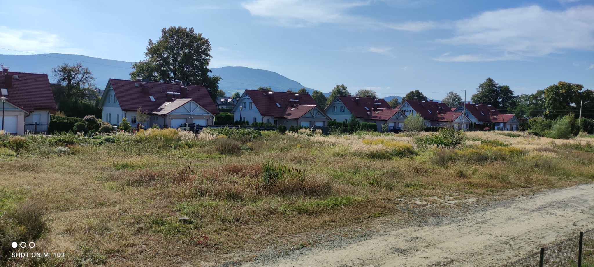 Rząd domów jednorodzinnych z czerwonymi dachami widziany zza zarośniętej łąki, na horyzoncie widoczne góry.