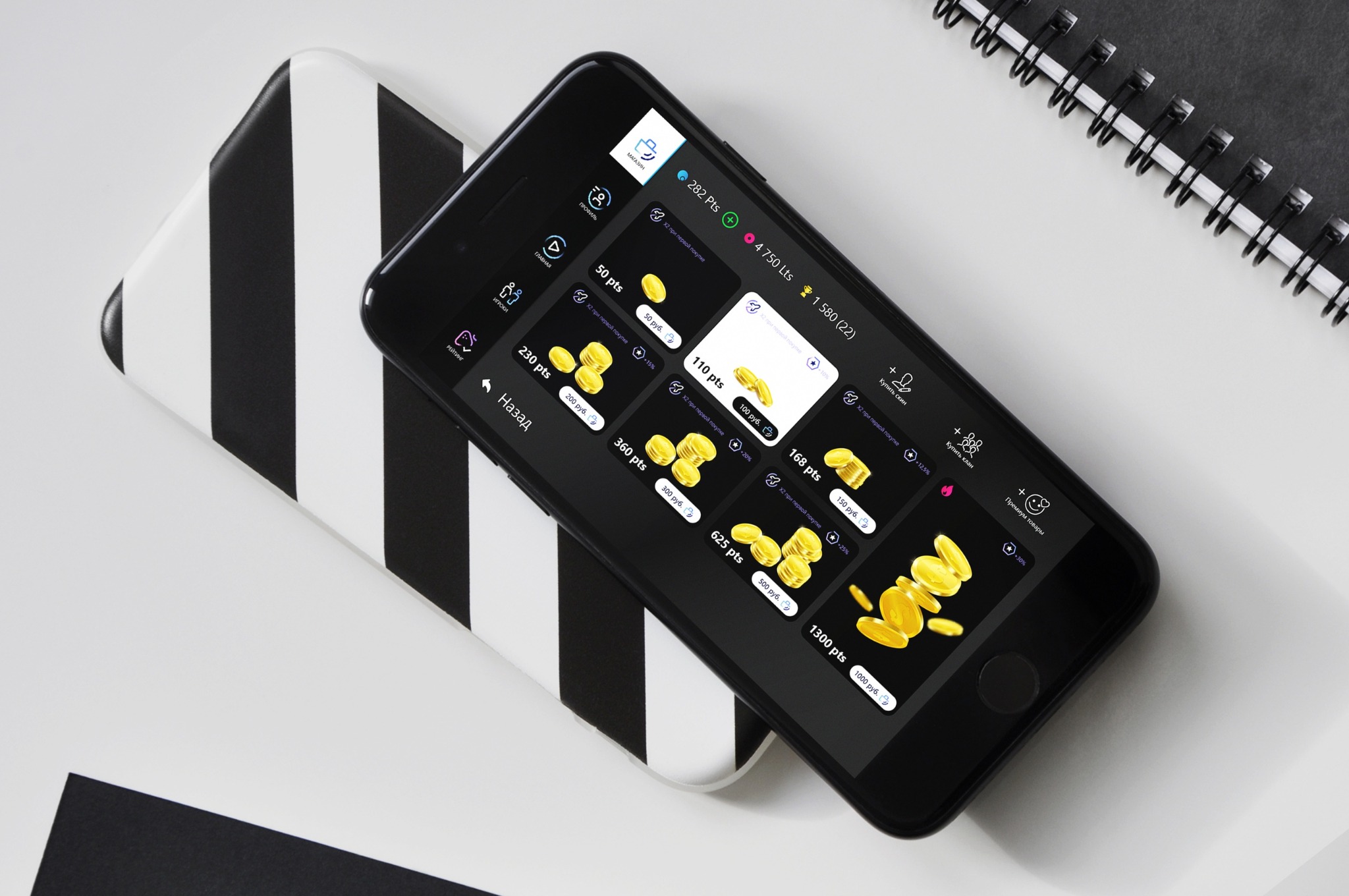 Ekran smartfona prezentujący interfejs aplikacji z motywem gromadzenia wirtualnych monet, umieszczony na podkładce w czarno-białe pasy obok czarnego notatnika.